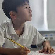 Student in a white shirt writing with a yellow pencil at a desk, looking toward the window.