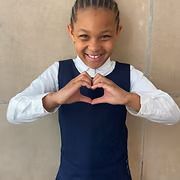 Smiling child in white shirt and navy vest making a heart shape with their hands against a beige wall