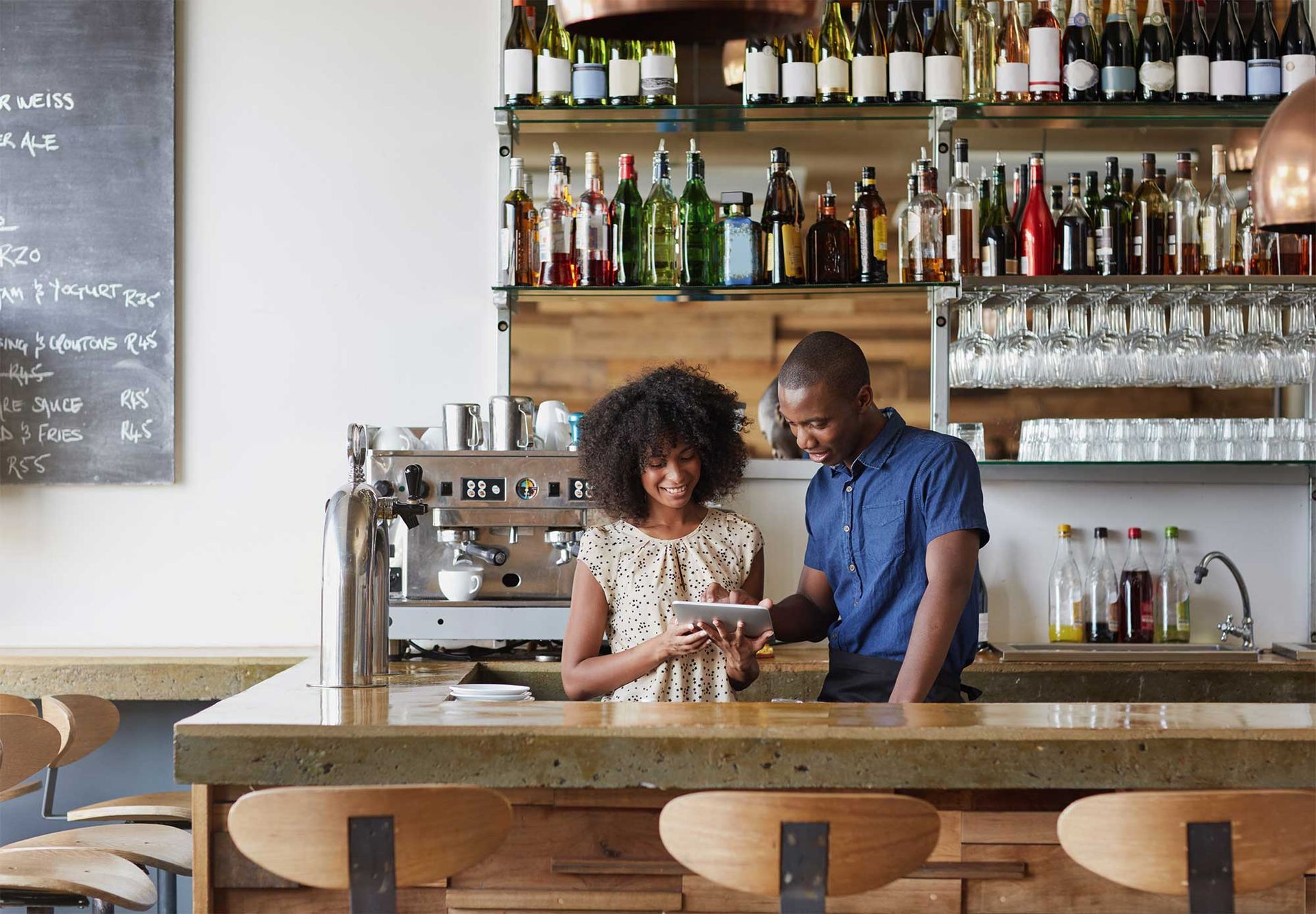 Two people, a man and woman, review a tablet at a bar counter with a liquor shelf in the background.