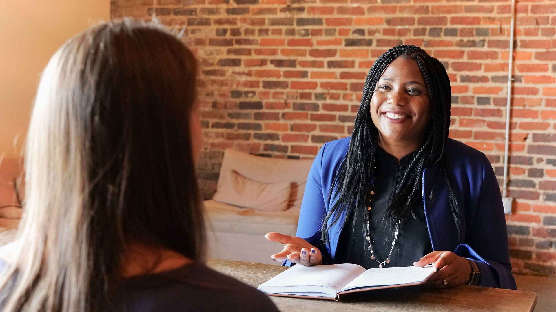 Woman with braids smiles while talking to another woman in front of a brick wall.