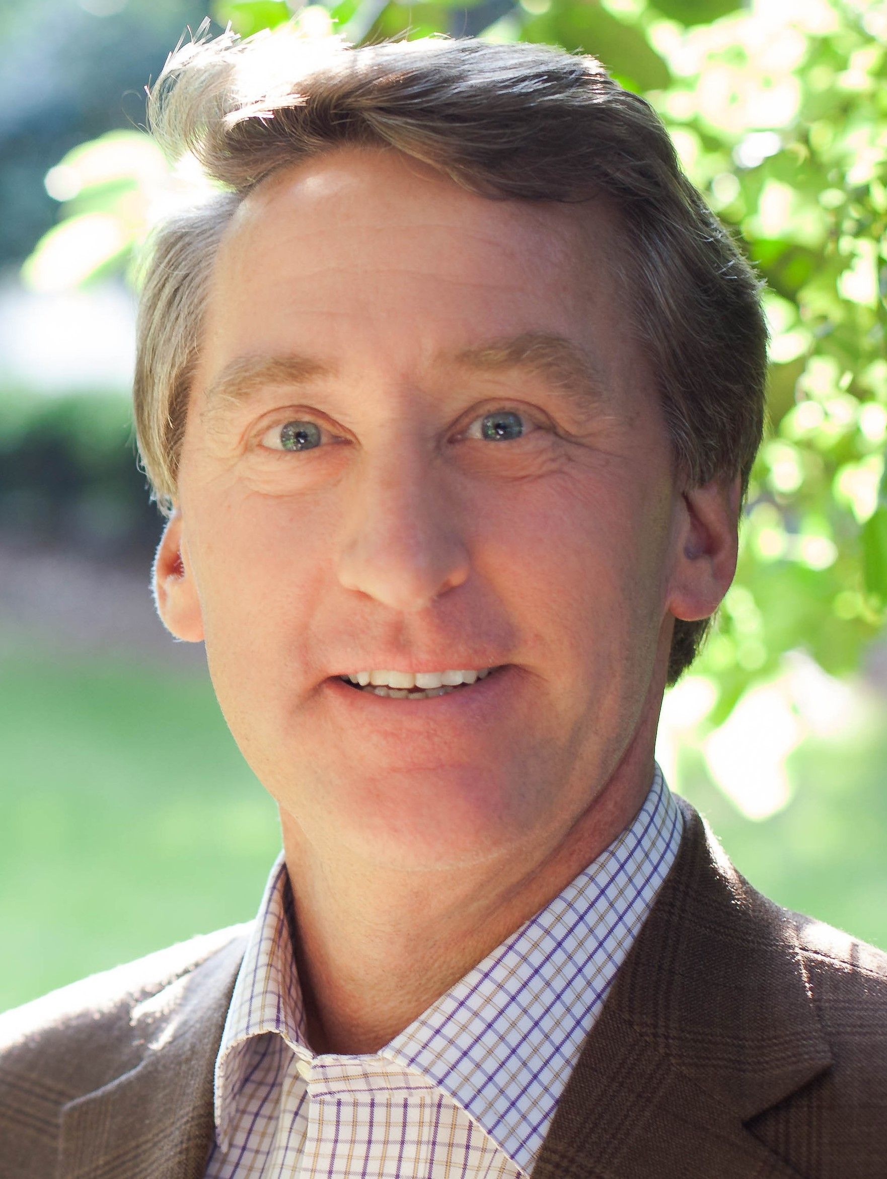 Man with light skin, smiling, wearing a brown suit jacket and a blue and white checkered shirt.