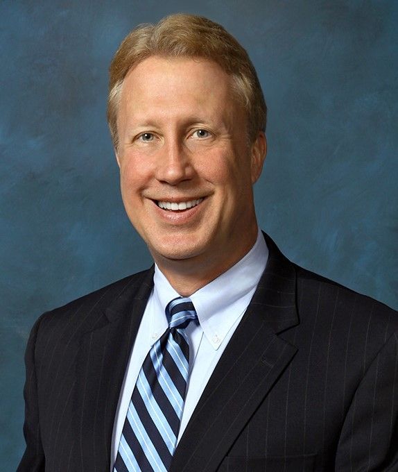Smiling, fair-skinned man in suit and tie against a blue backdrop.