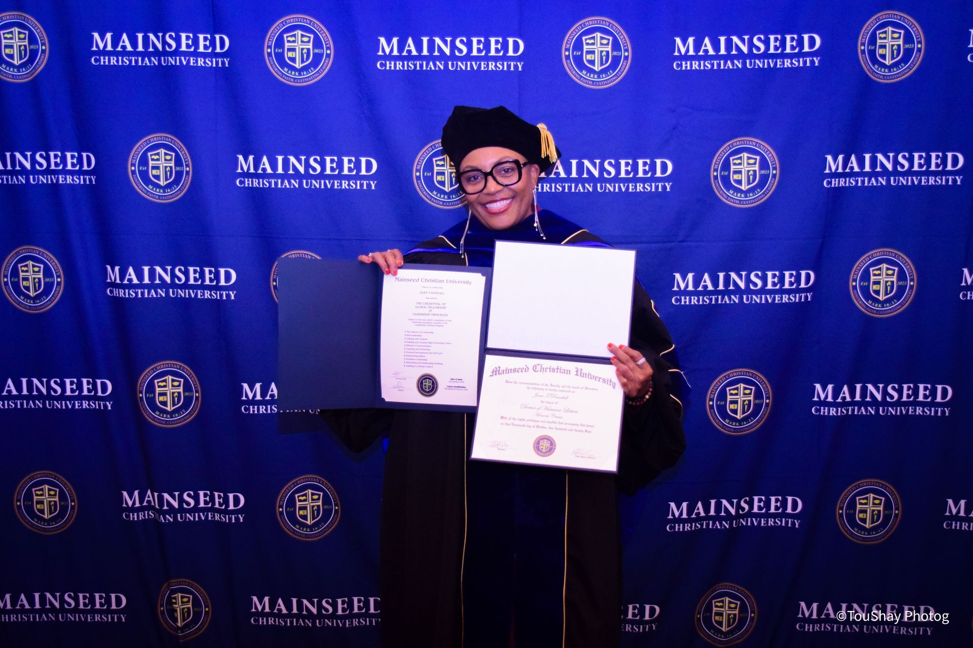 A person in cap and gown holds a diploma, posing in front of a university backdrop.