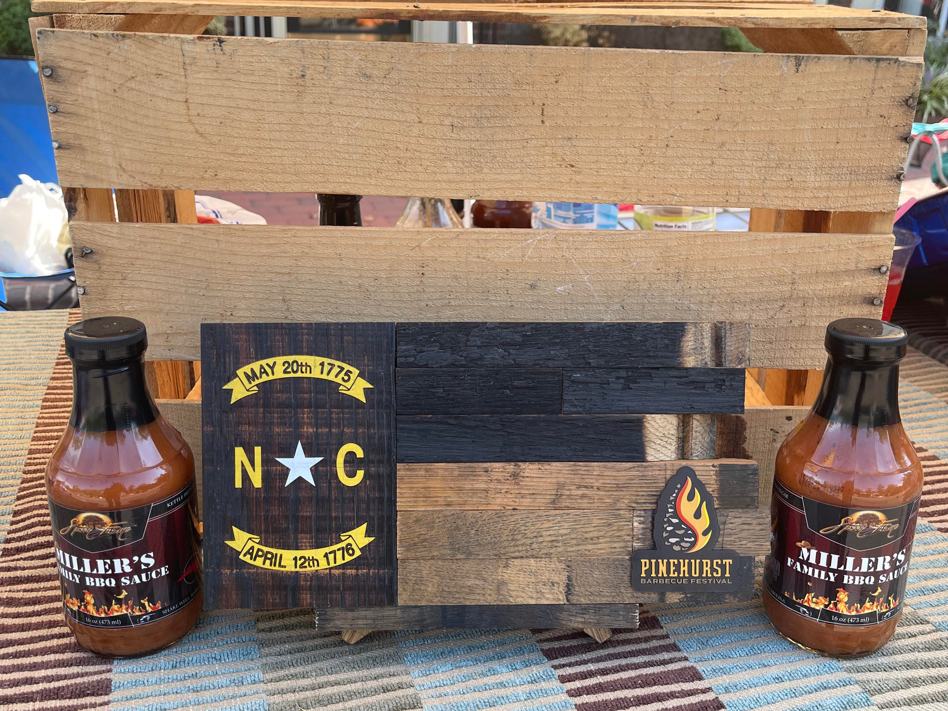 Two bottles of BBQ sauce flank a wooden sign with the North Carolina flag; all in front of a wooden pallet.