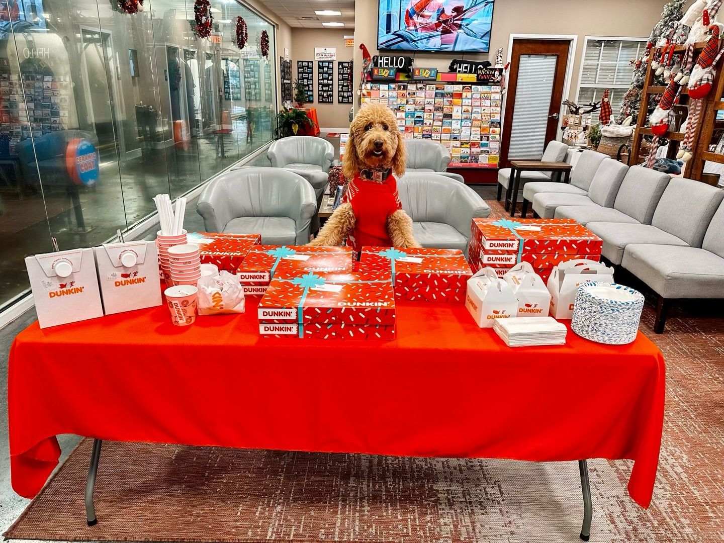Dog in red shirt sits atop a table covered with red boxes and bags. Indoor setting with holiday decorations.