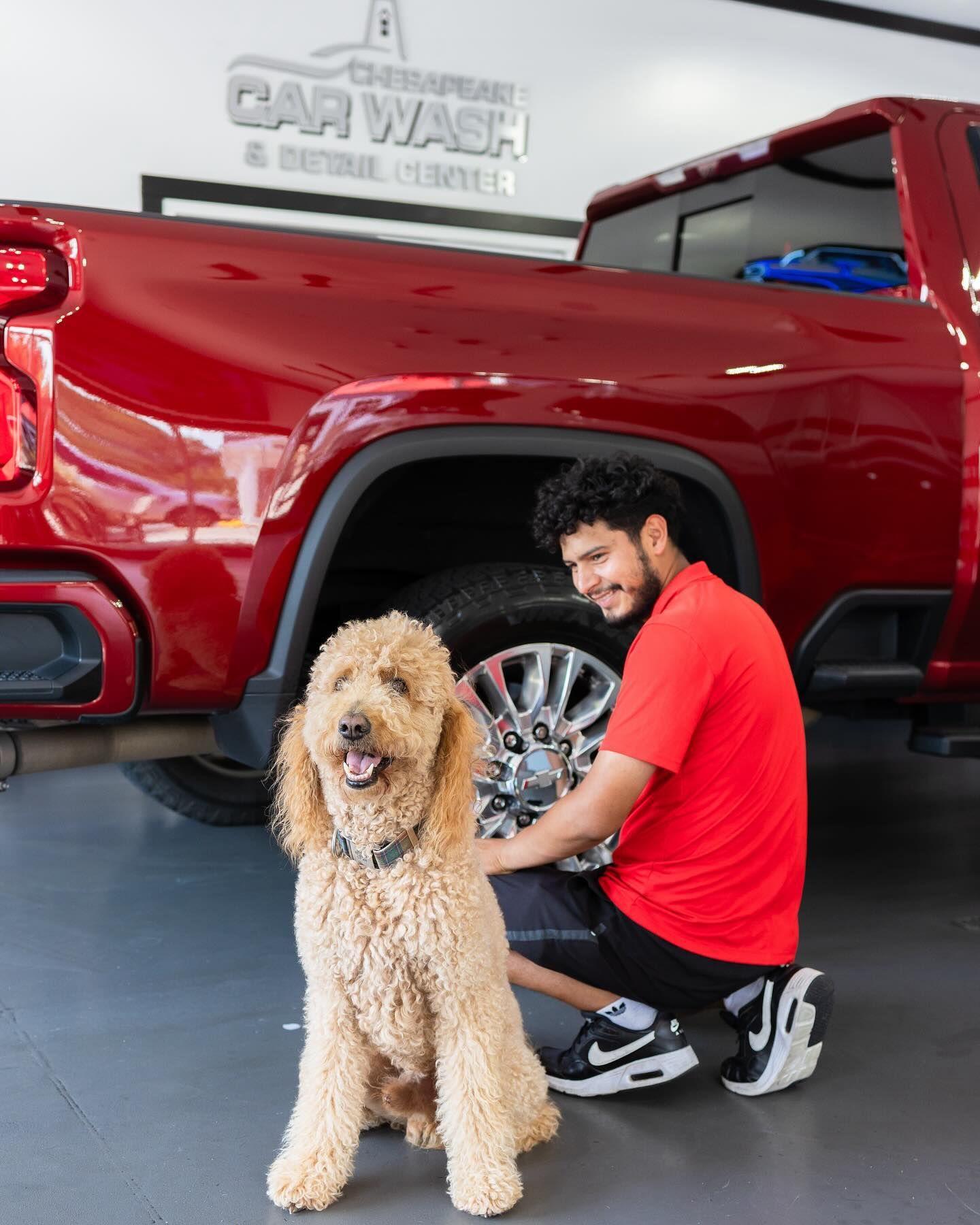 Man in red shirt and a golden doodle dog by a red truck tire, car wash setting.