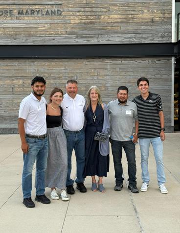 Six people posing together outside a building with a wooden facade.