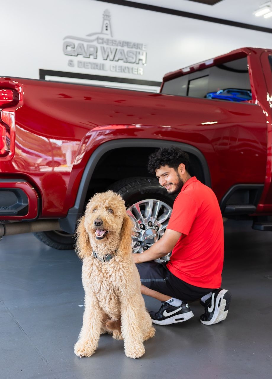 Man in red shirt crouches beside a fluffy tan dog in front of a red pickup truck at a car wash