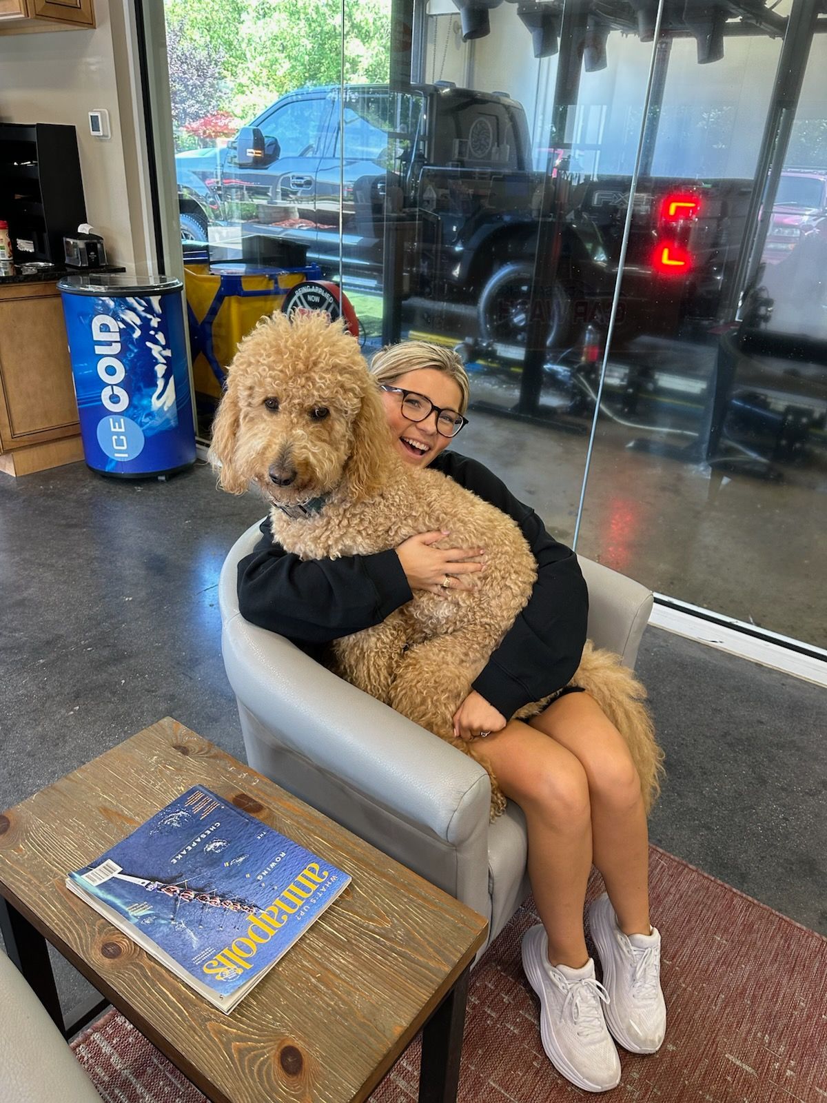 Person hugging a fluffy tan dog in a chair by a glass storefront, with a blue book on the table.