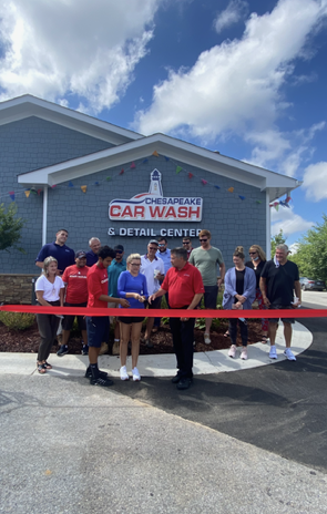 Group stands at a car wash ribbon-cutting in front of a blue building under a cloudy sky.