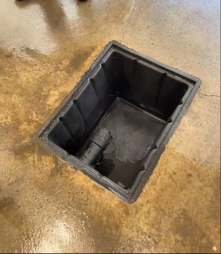 Trappe d'accès au sol révélant une fosse utilitaire noire avec un tuyau à l'intérieur, sur un sol en béton beige.