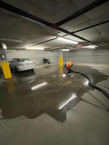Une personne vêtue d'orange travaille avec un tuyau d'arrosage dans un parking inondé, à côté d'une voiture blanche.