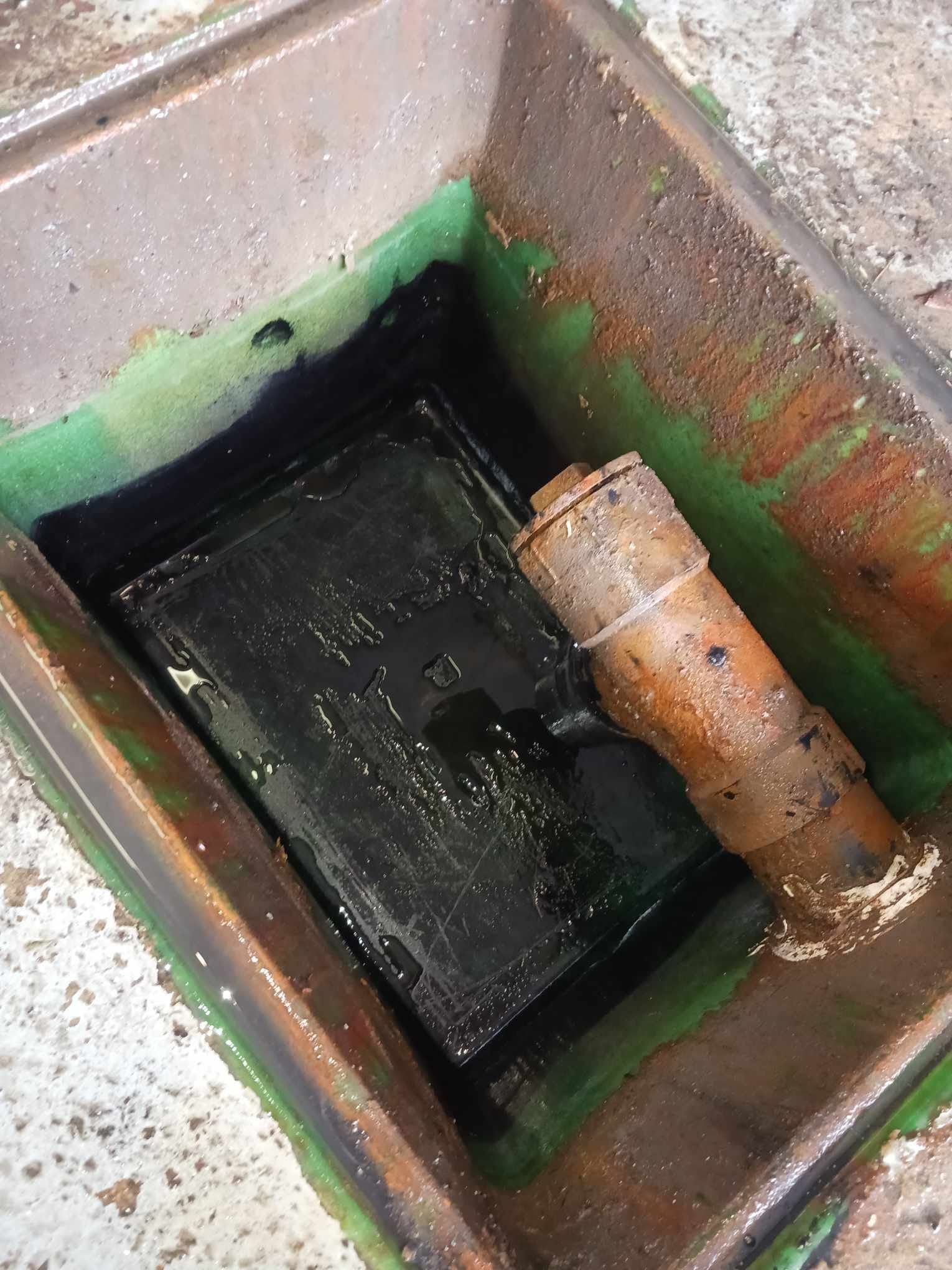 Puits de service ouvert avec couvercle noir et tuyau rouillé à l'intérieur du puits en béton.