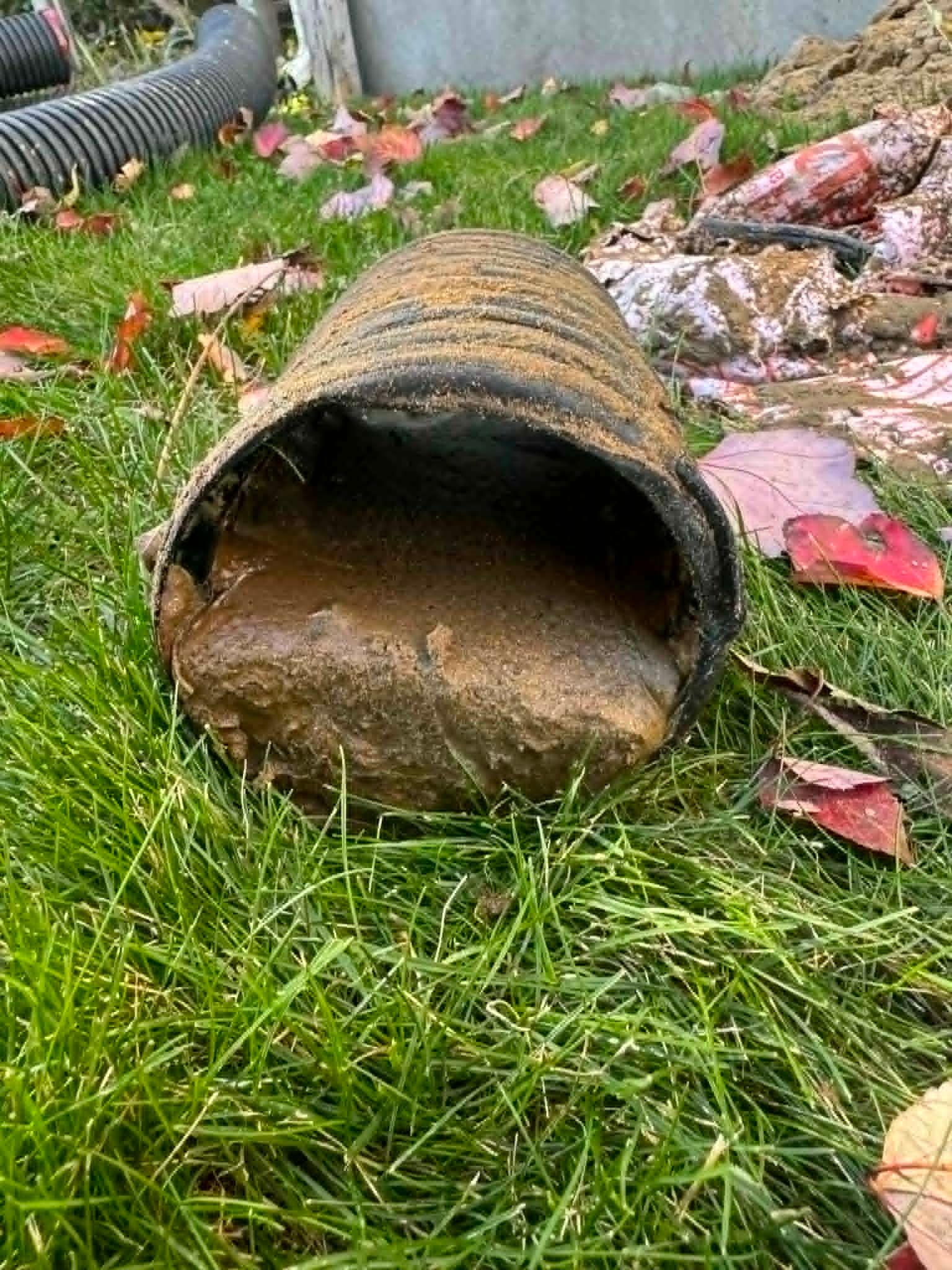 Un tuyau noir effondré gît sur l'herbe, à côté de débris et de boue éparpillés.