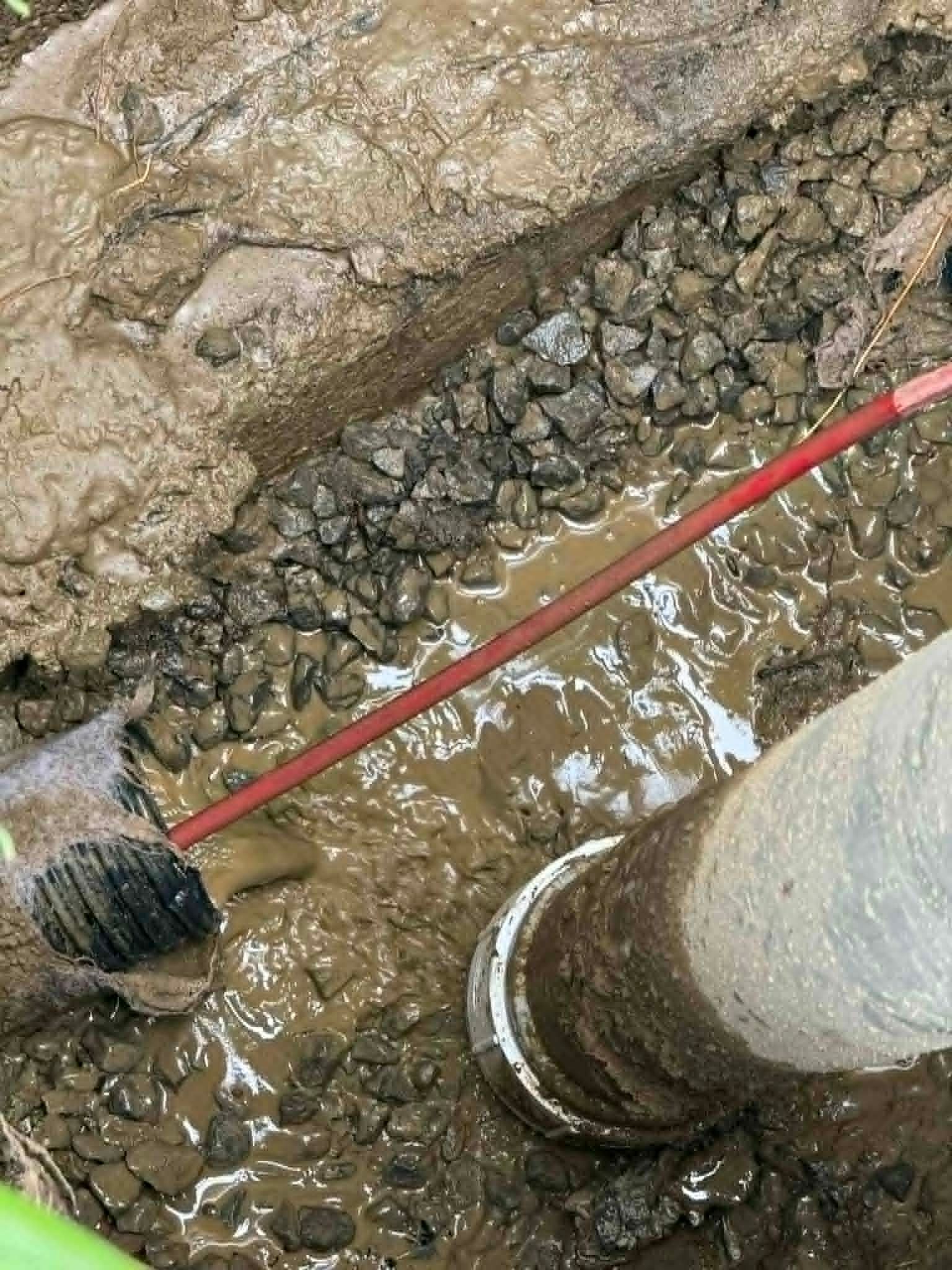Un tuyau rouge longe une tranchée boueuse, avec un tuyau humide et du gravier sous un sol rocailleux.