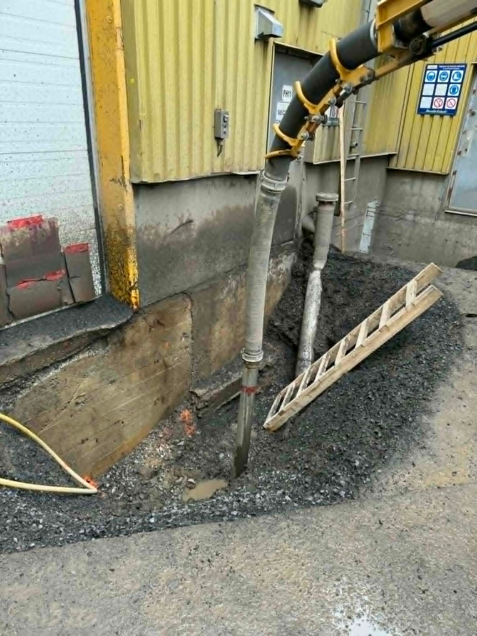 Tranchée technique en béton à côté d'un bâtiment jaune, avec des tuyaux apparents et du gravier au fond.