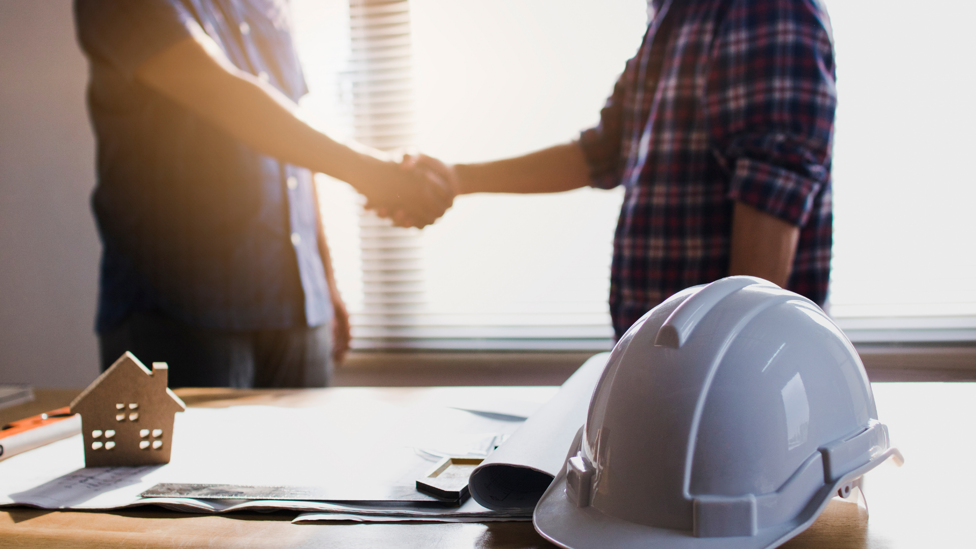 Two people shaking hands across a desk with a white hard hat in the foreground in a bright office