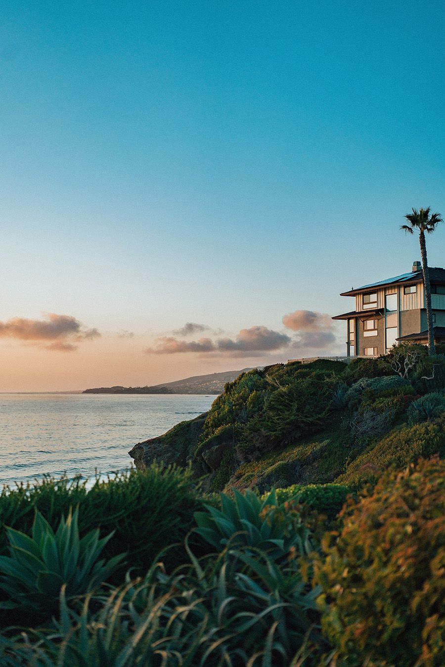 Coastal house at sunset, overlooking the ocean. Blue sky, golden light. Green foliage in the foreground.