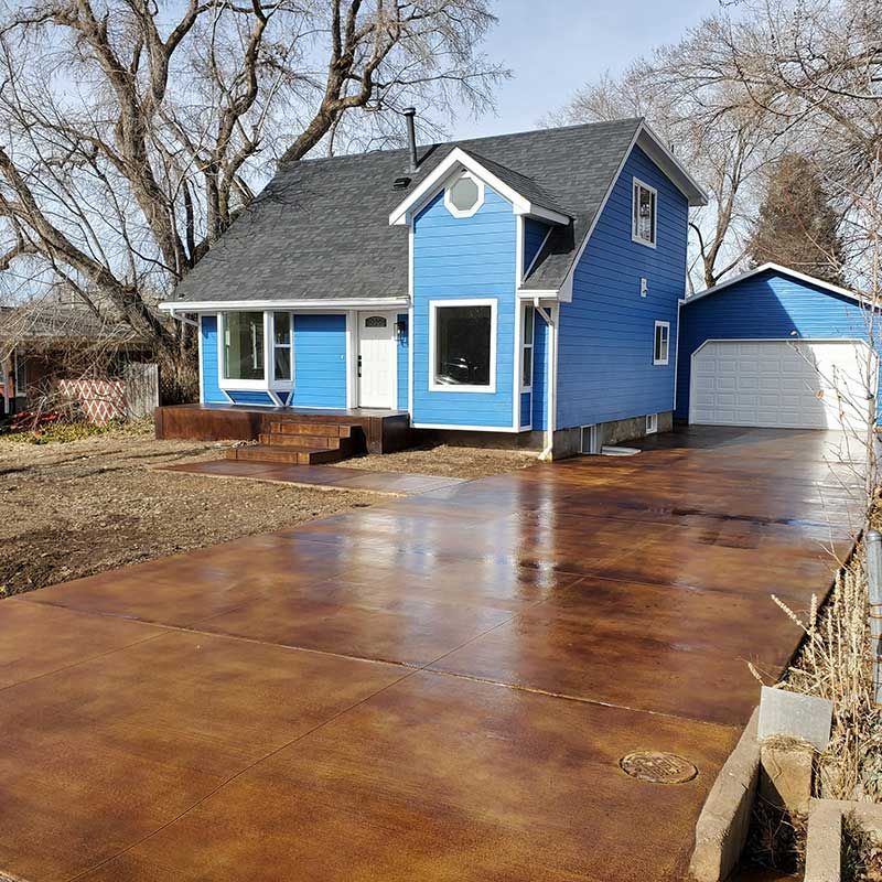 Blue house with a small porch and a long wet concrete driveway beside a detached garage