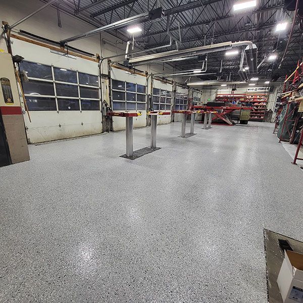 Empty industrial workshop with concrete floor, overhead lights, and garage doors along the left wall.