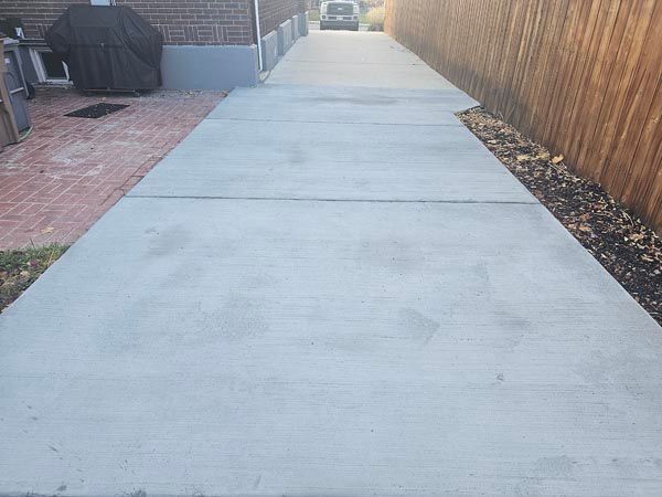 Concrete backyard walkway beside brick patio and wooden fence, with outdoor grill on the left.