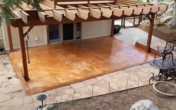 Wooden pergola over a wet stone patio beside a house, with metal chairs and table on the right.