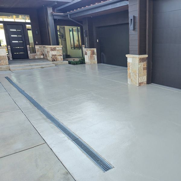 Residential driveway with garage doors, stone columns, and a long drainage grate down the center.