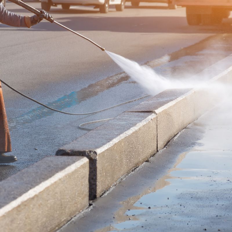Pressure washing a curb and pavement, spraying water across a wet roadway