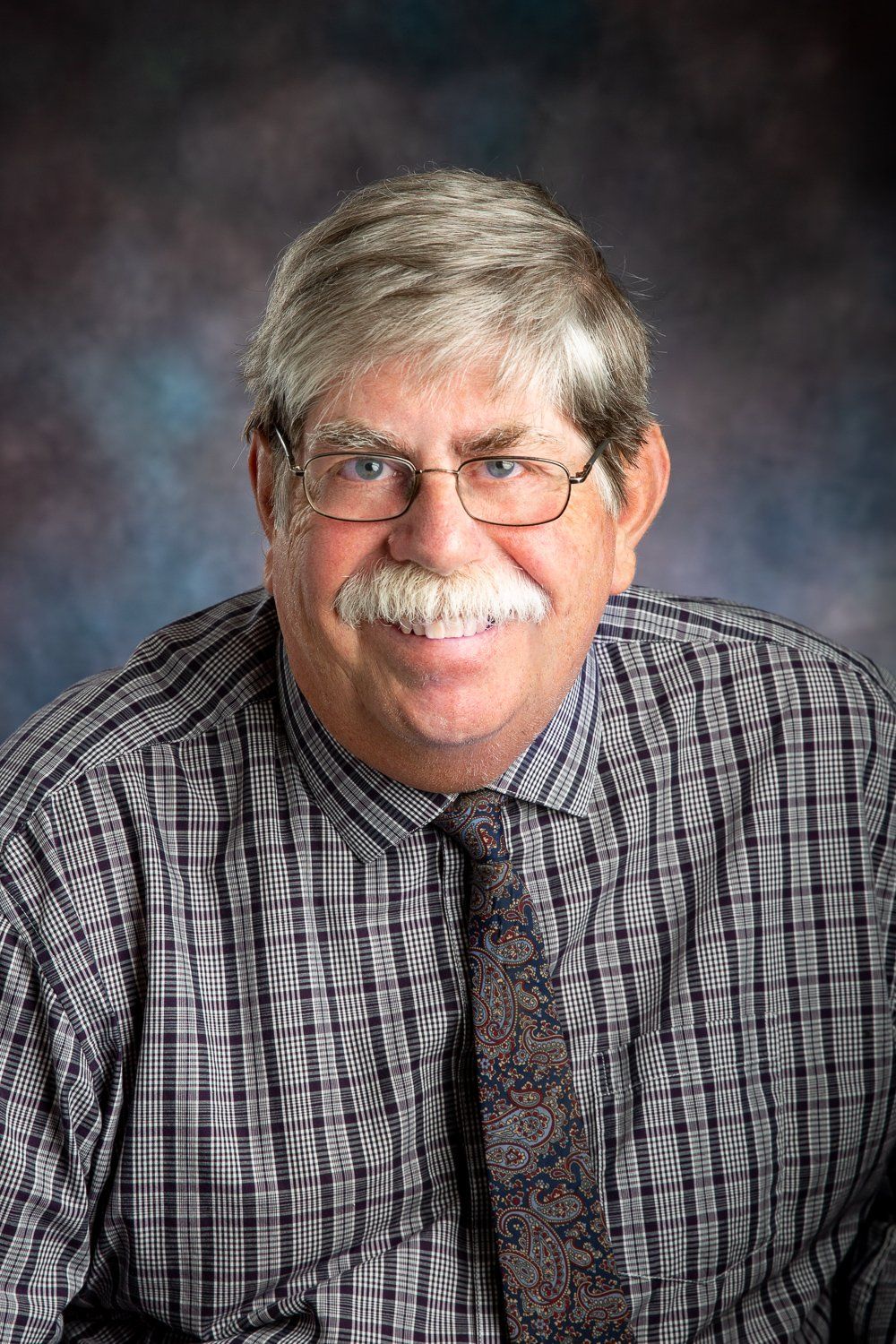 A man with glasses and a mustache is wearing a plaid shirt and tie.