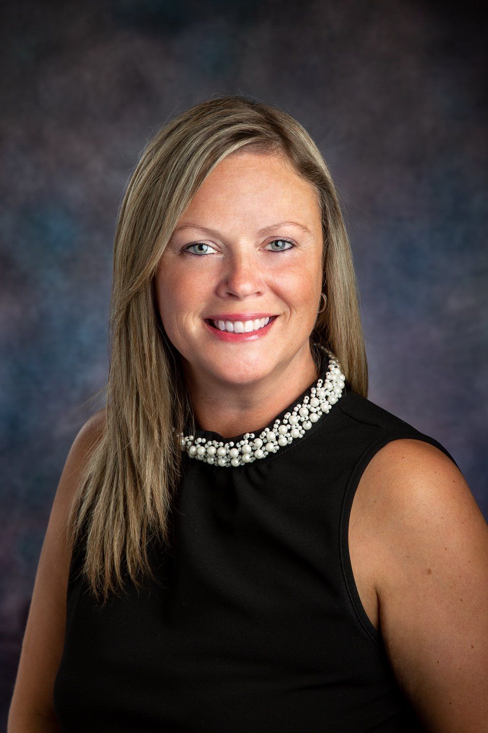 A woman in a black dress with a pearl collar is smiling for the camera.