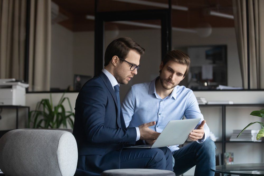 Two men looking at a laptop in an office. One in a suit, the other in a button-down shirt.