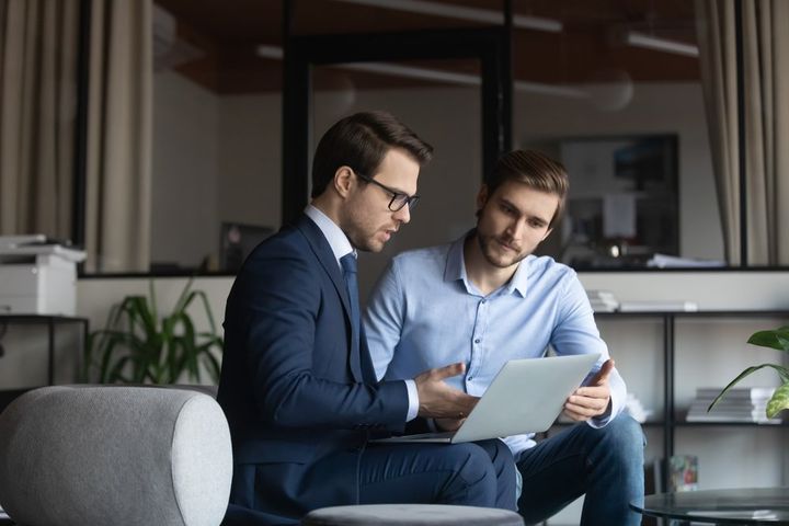 Two men in an office looking at a laptop. One in a suit, one in a blue shirt.