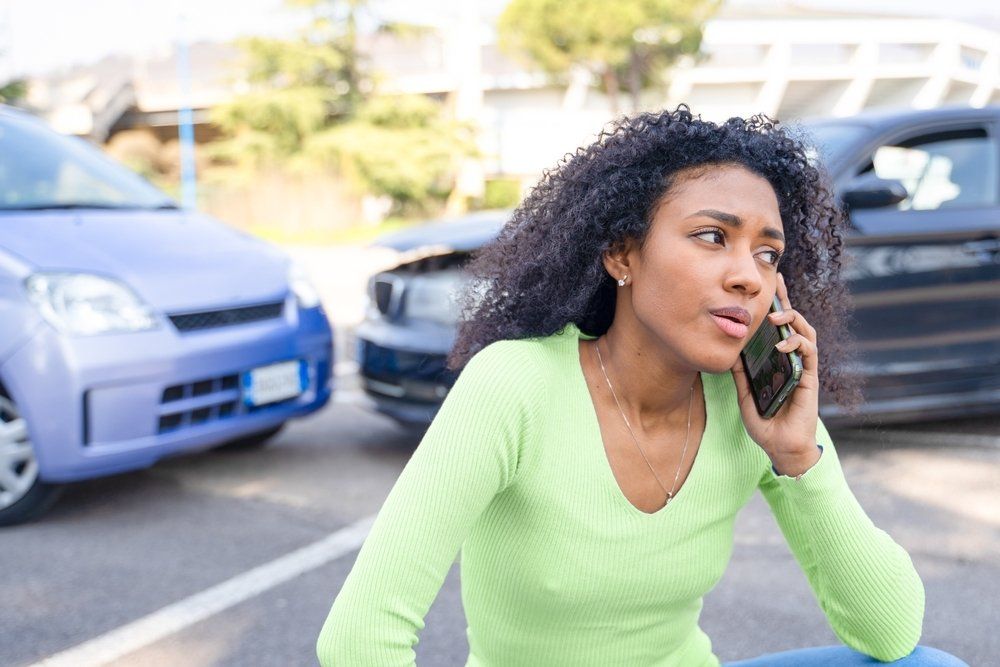 Woman on phone at car accident scene, looking concerned. Two damaged cars in background.