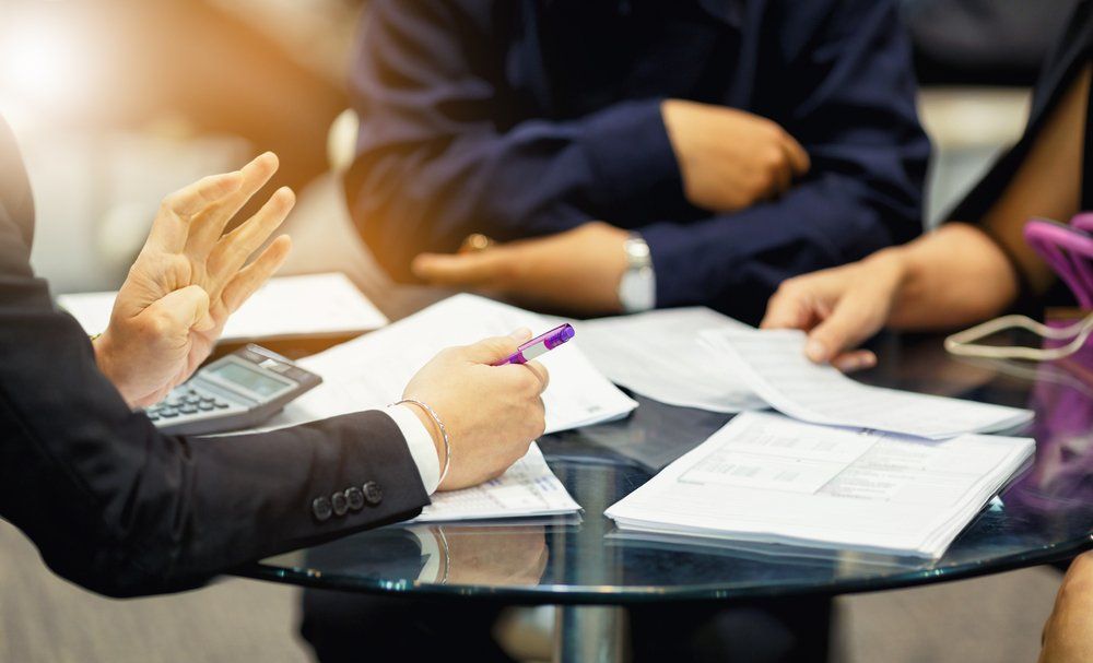 Businesspeople in suits around a table, documents and a calculator present, discussing and gesturing.