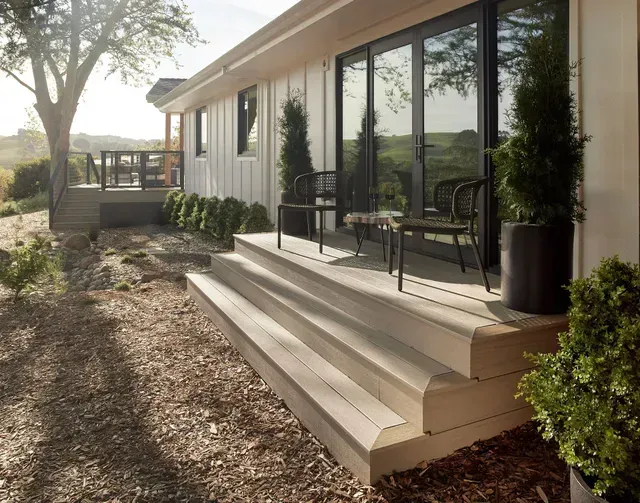 Exterior of a white house with wooden steps leading to a deck and glass doors. Two chairs sit on the deck.