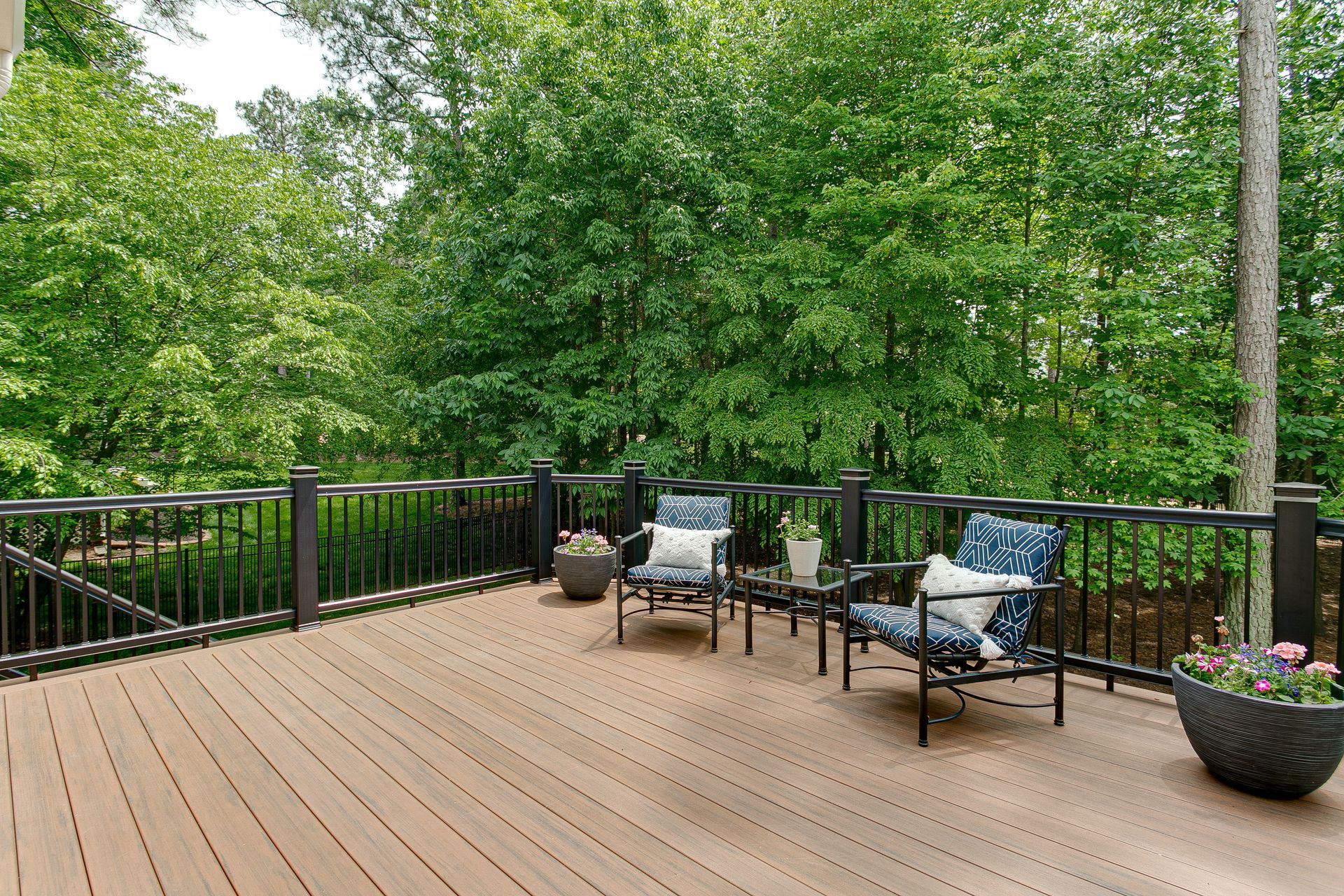 Wooden deck with two patterned armchairs, a small table, and potted plants, surrounded by trees.