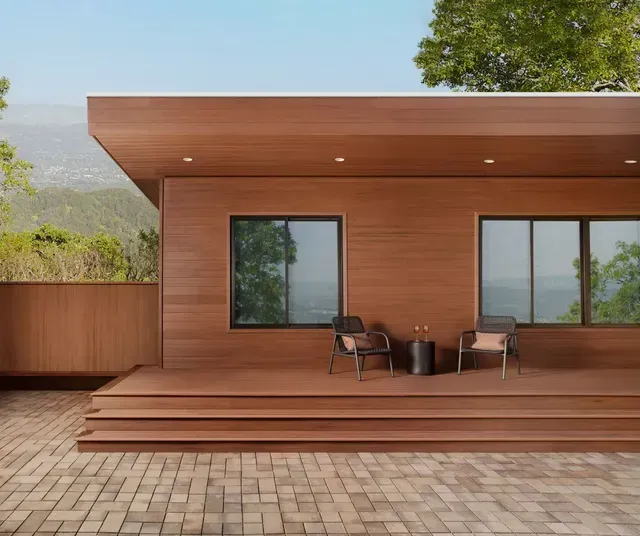 Wooden modern house with deck, chairs, and brick patio. Mountain backdrop.