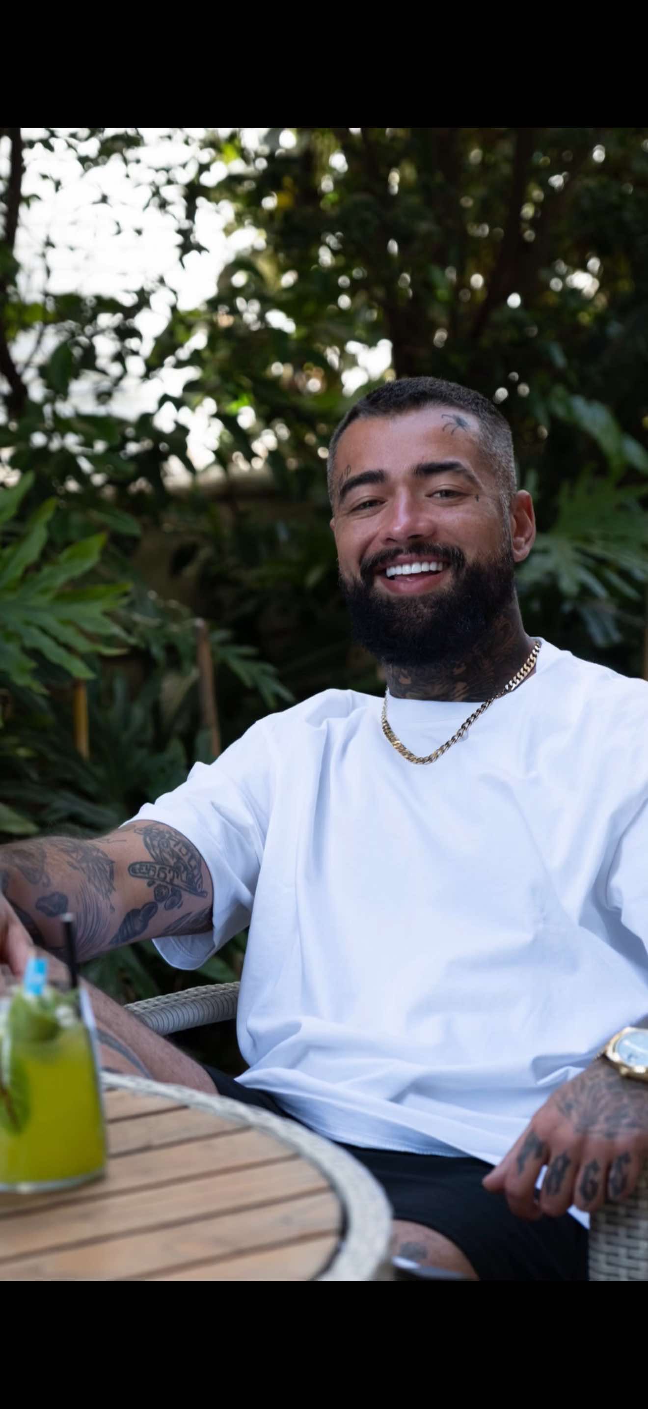 A man with tattoos smiles, sitting outdoors with a drink. He wears a white t-shirt, and a gold chain.