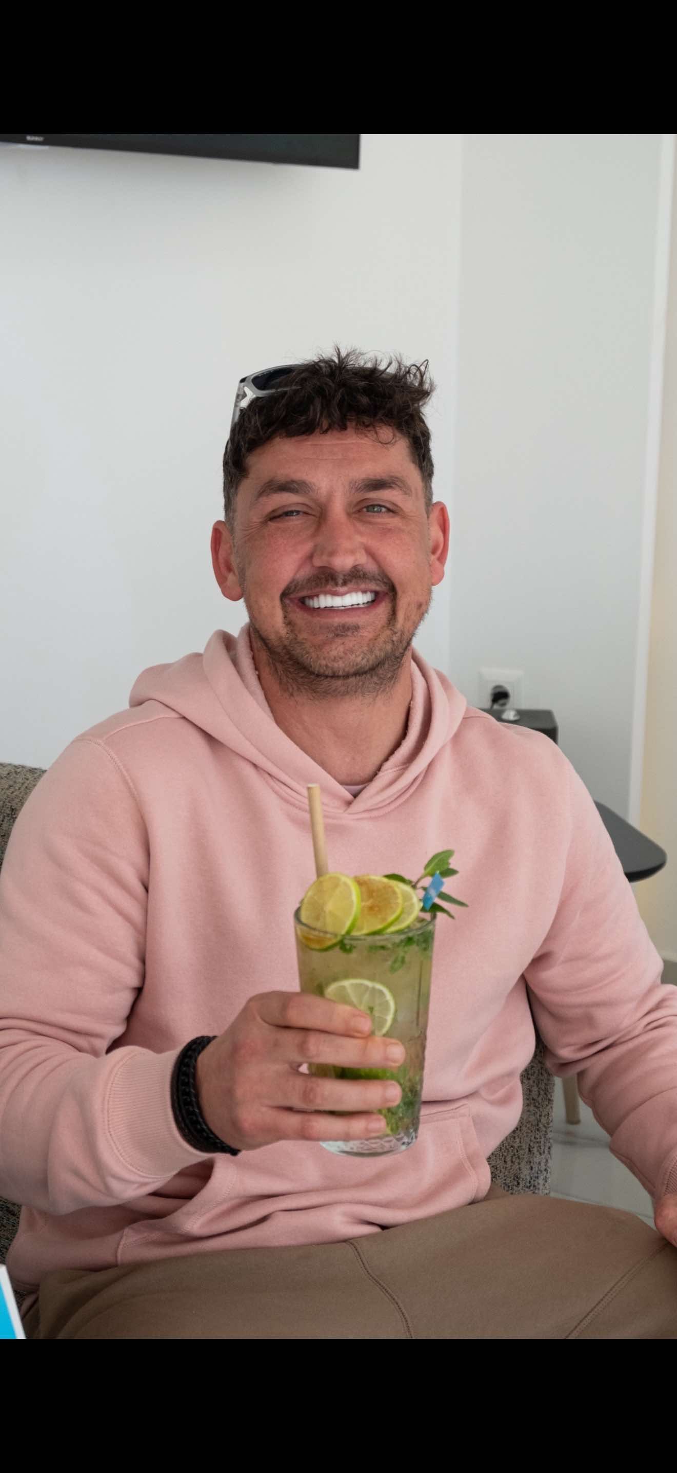 A smiling person holds a cocktail with lime and mint in a clear glass. They wear a pink hoodie.