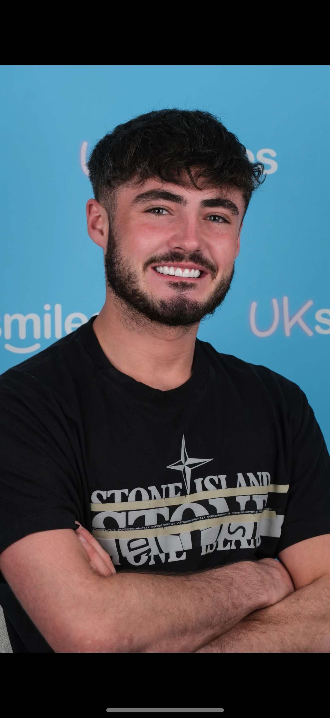 Man smiling, arms crossed, wearing a black Stone Island shirt, against a blue backdrop.