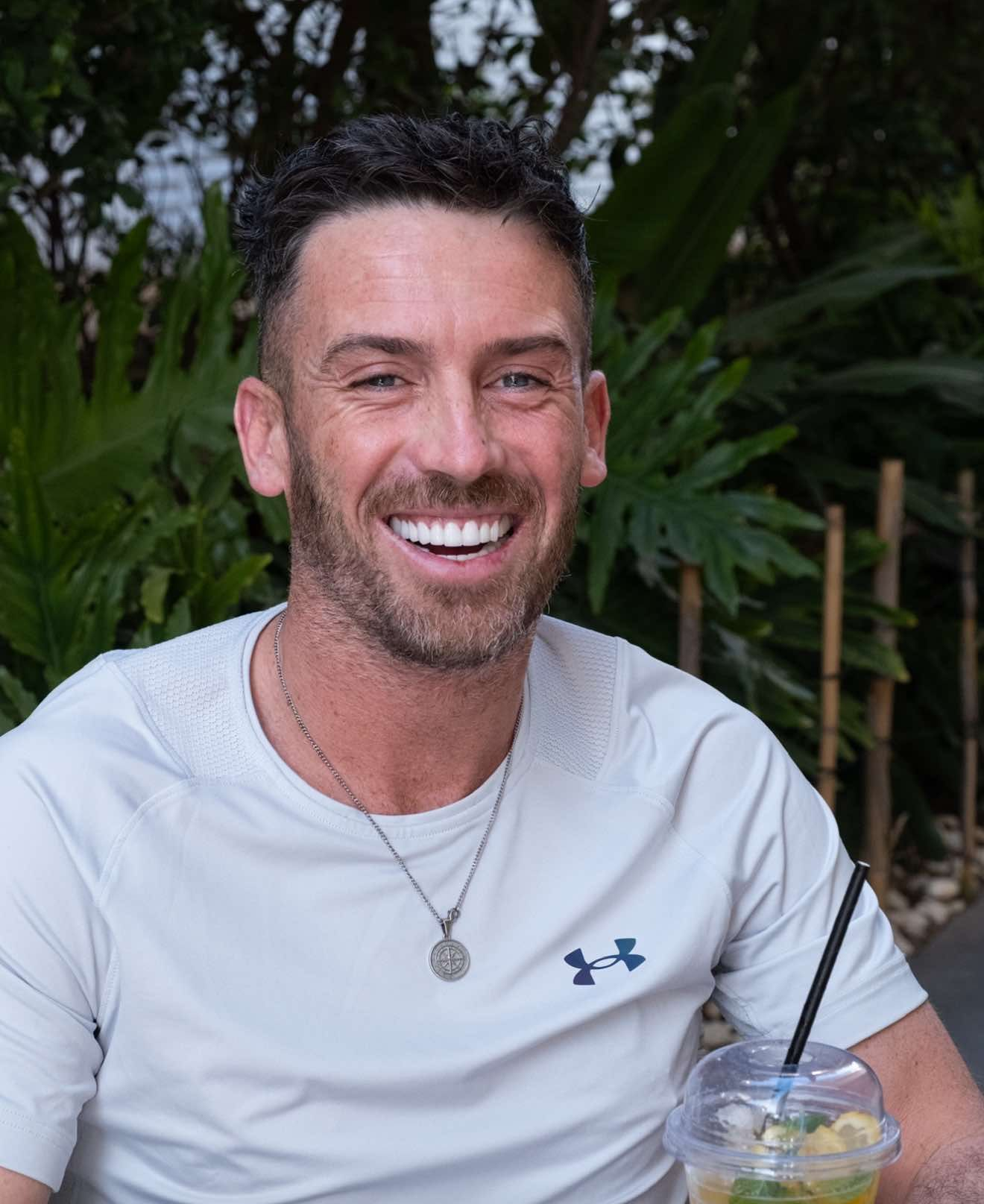 Man with a smile wearing a light gray shirt, necklace, sitting outdoors with a drink.