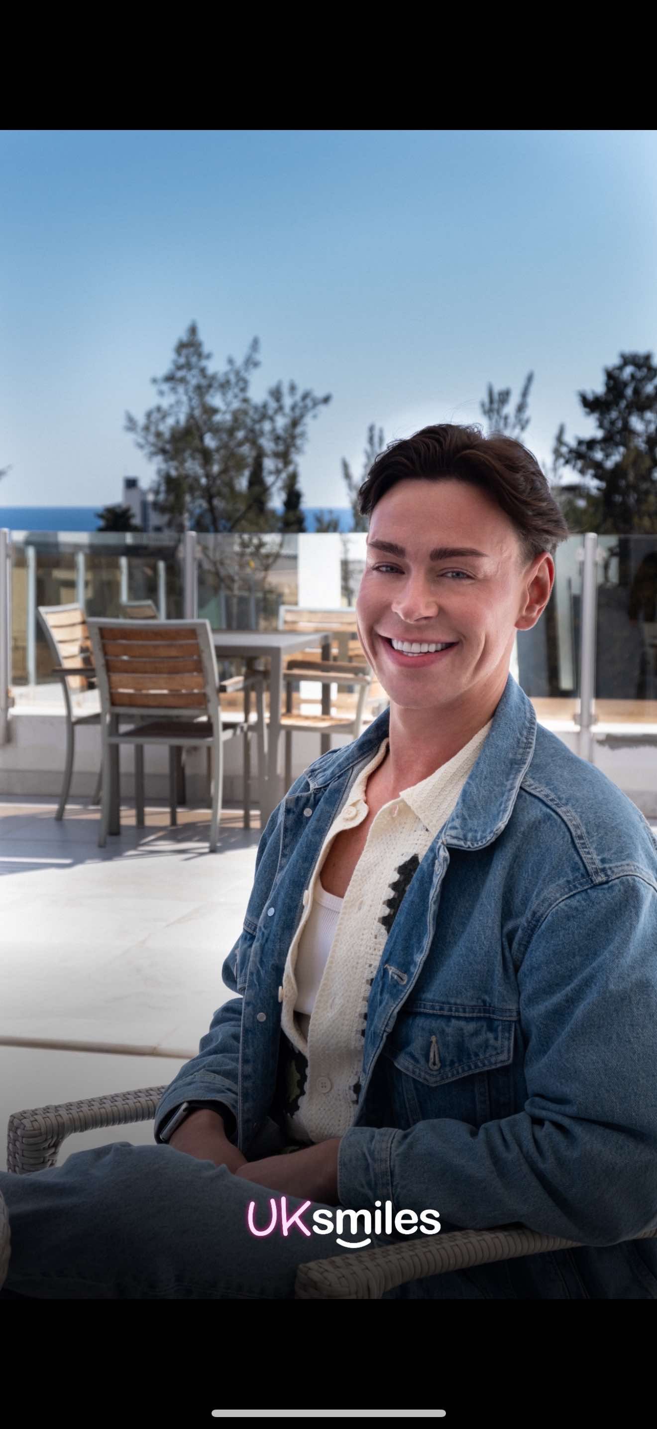 Woman in denim jacket smiles outdoors on a sunny patio.