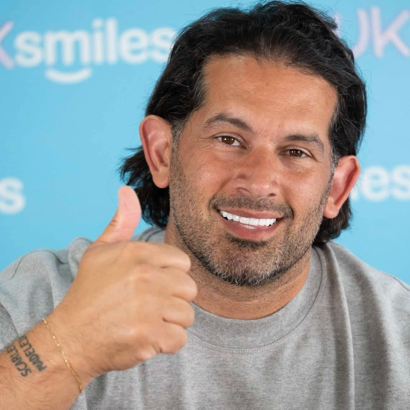 Man with dark hair and white teeth, giving a thumbs-up. Blue background.