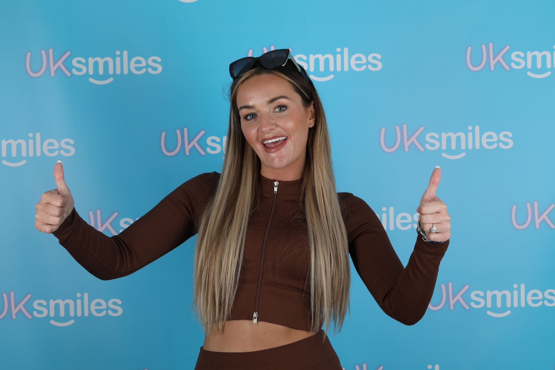 Woman in brown outfit gives thumbs up in front of a blue 