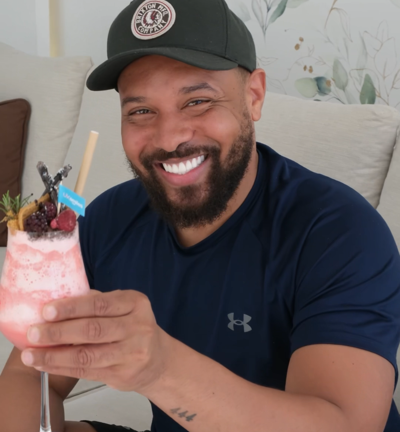 Man smiling, holding a pink cocktail with fruit garnish, wearing a dark cap and shirt.
