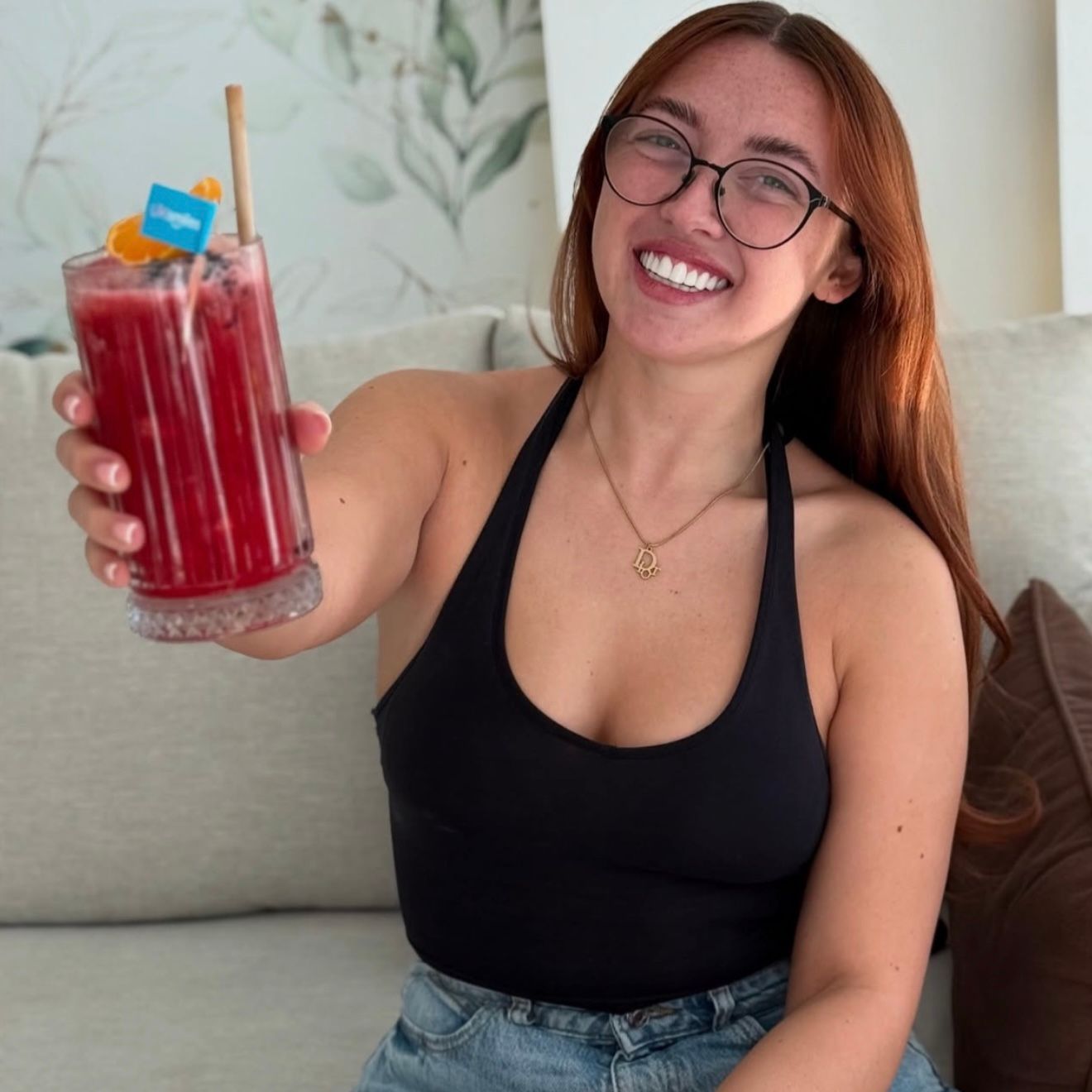 Woman in black halter top holds a red drink with orange slice and straw, smiling, indoors.