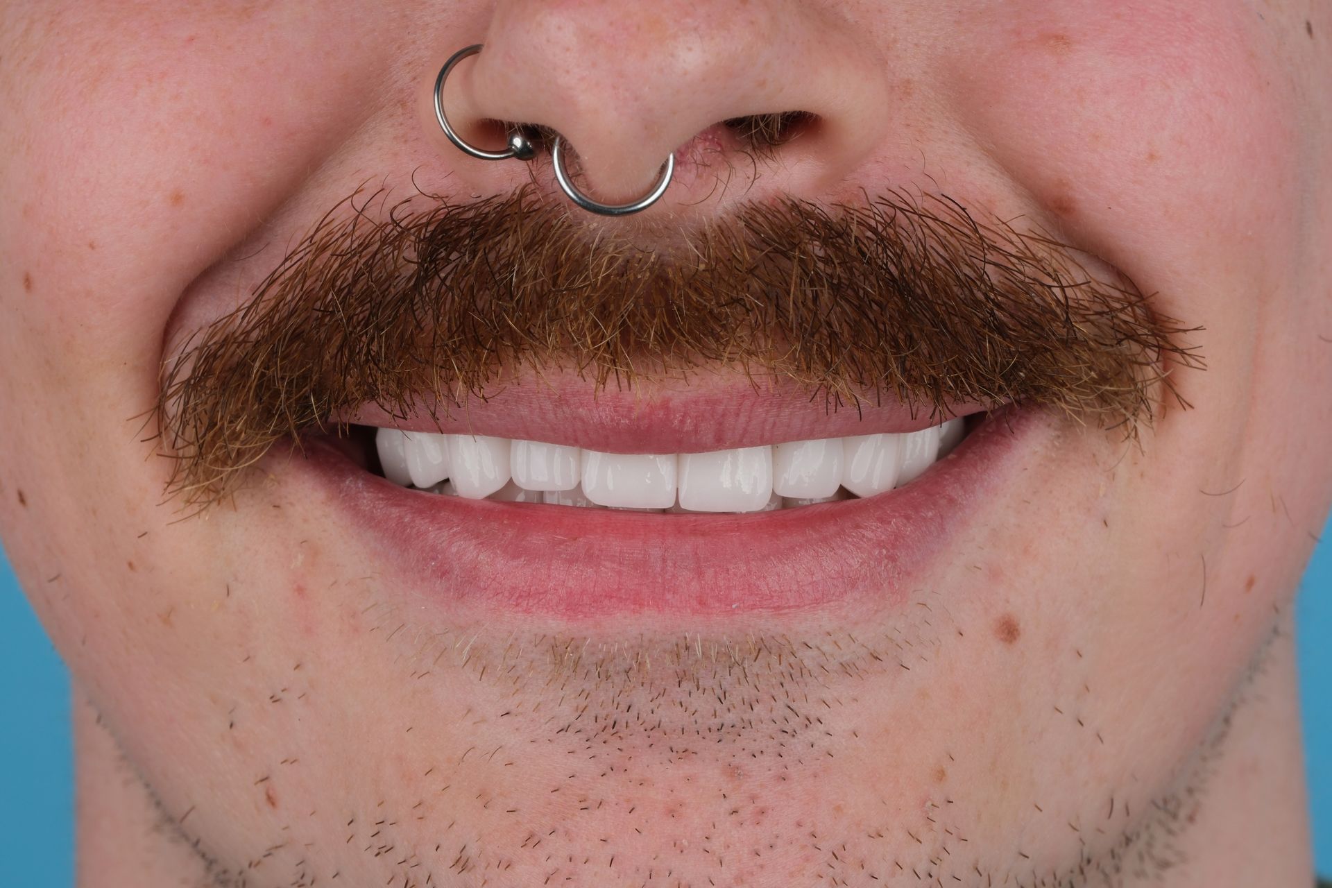 Close-up of a person smiling with a large brown mustache, septum piercing, and bright white teeth.
