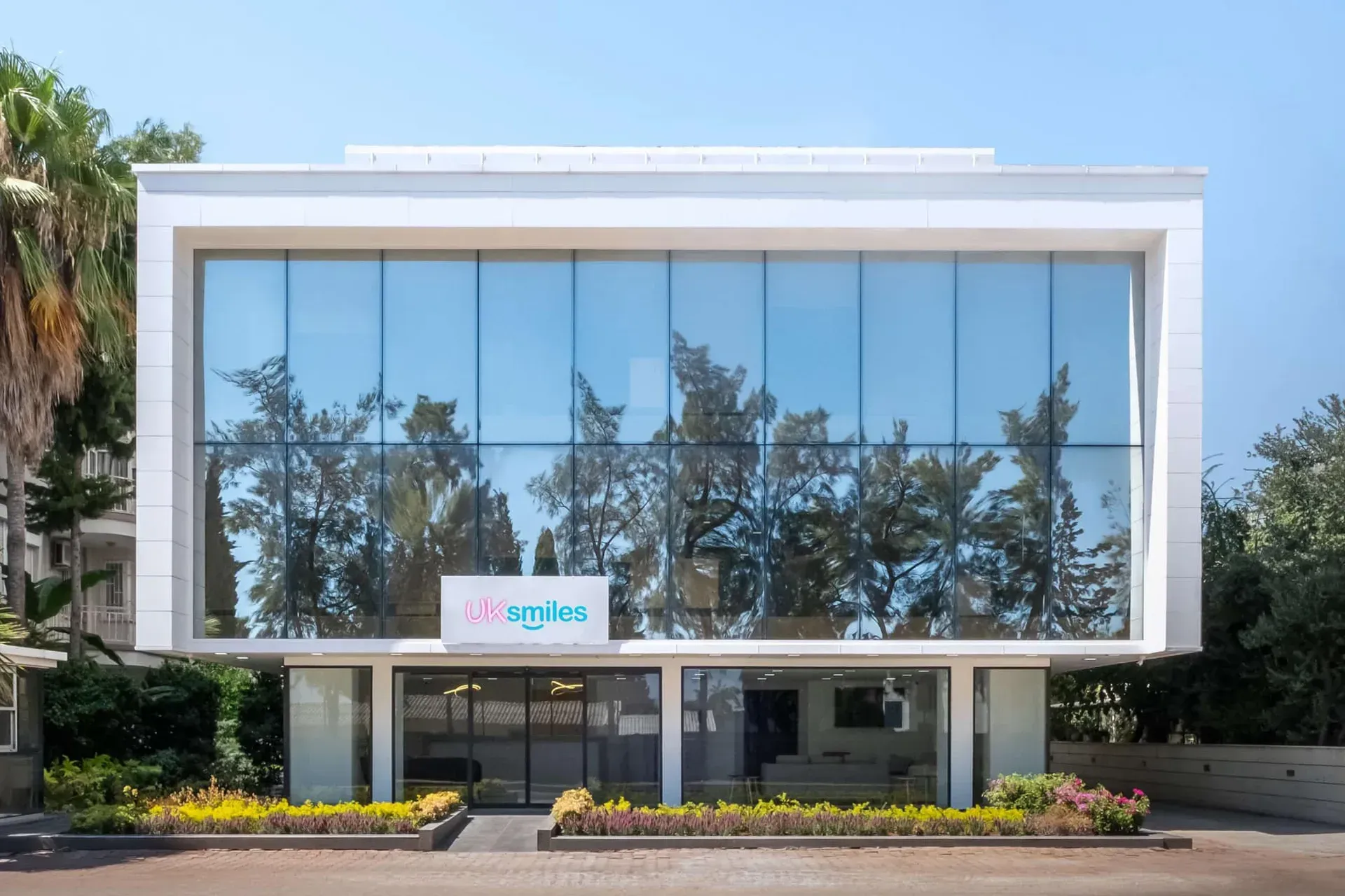 Modern white building with large glass windows reflecting trees and sky; logo 