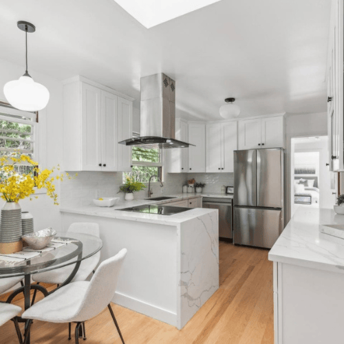Bright white modern kitchen with stainless steel fridge, island, pendant lights, and a small dining table with flowers.