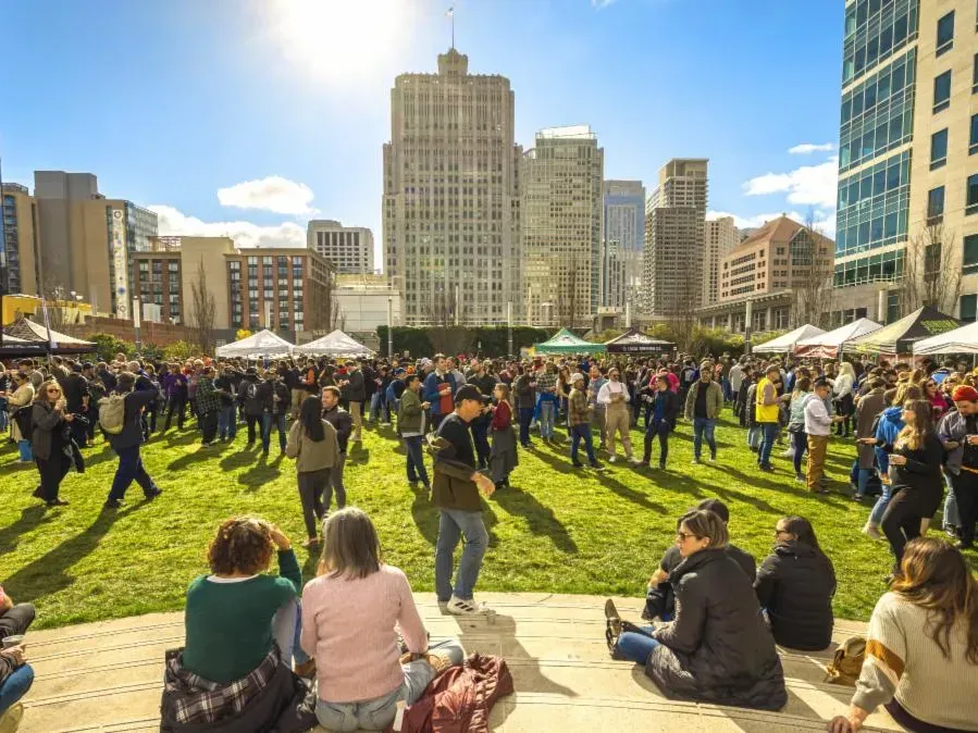 Crowd gathered in a sunny city park with tents, seated people on grass, and tall buildings in the background